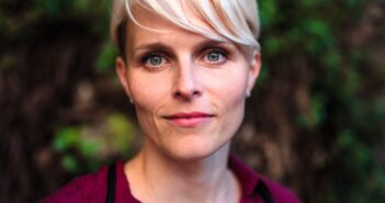 A woman with short blonde hair and blue eyes looks directly at the camera, wearing a burgundy shirt, with a blurred outdoor background of greenery.
