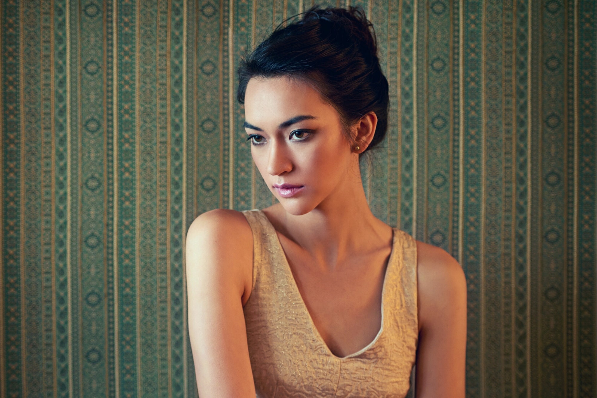 A young woman with dark hair in an updo, wearing a sleeveless beige dress, stands against a patterned green wallpaper, looking to her left with a contemplative expression.
