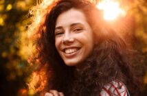 A woman with long, curly dark hair smiles warmly at the camera. She is outdoors with sunlight shining behind her, creating a golden, glowing effect around her head.