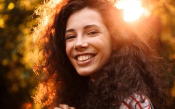 A woman with long, curly dark hair smiles warmly at the camera. She is outdoors with sunlight shining behind her, creating a golden, glowing effect around her head.