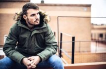 A young man with short hair and a beard sits outdoors on a ledge, wearing a green parka with a fur-lined hood and blue jeans, looking thoughtfully to the side. Urban buildings are blurred in the background.