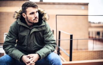 A young man with short hair and a beard sits outdoors on a ledge, wearing a green parka with a fur-lined hood and blue jeans, looking thoughtfully to the side. Urban buildings are blurred in the background.