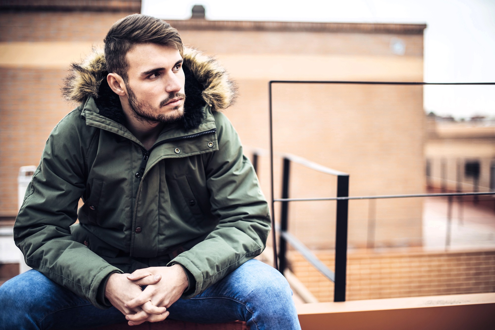 A young man with short hair and a beard sits outdoors on a ledge, wearing a green parka with a fur-lined hood and blue jeans, looking thoughtfully to the side. Urban buildings are blurred in the background.
