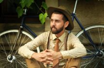 A bearded man in a polka-dot shirt, tie, suspenders, and wide-brimmed hat sits cross-legged outdoors, hands clasped, with a vintage bicycle and greenery in the background.