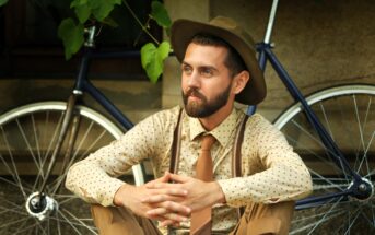 A bearded man in a polka-dot shirt, tie, suspenders, and wide-brimmed hat sits cross-legged outdoors, hands clasped, with a vintage bicycle and greenery in the background.