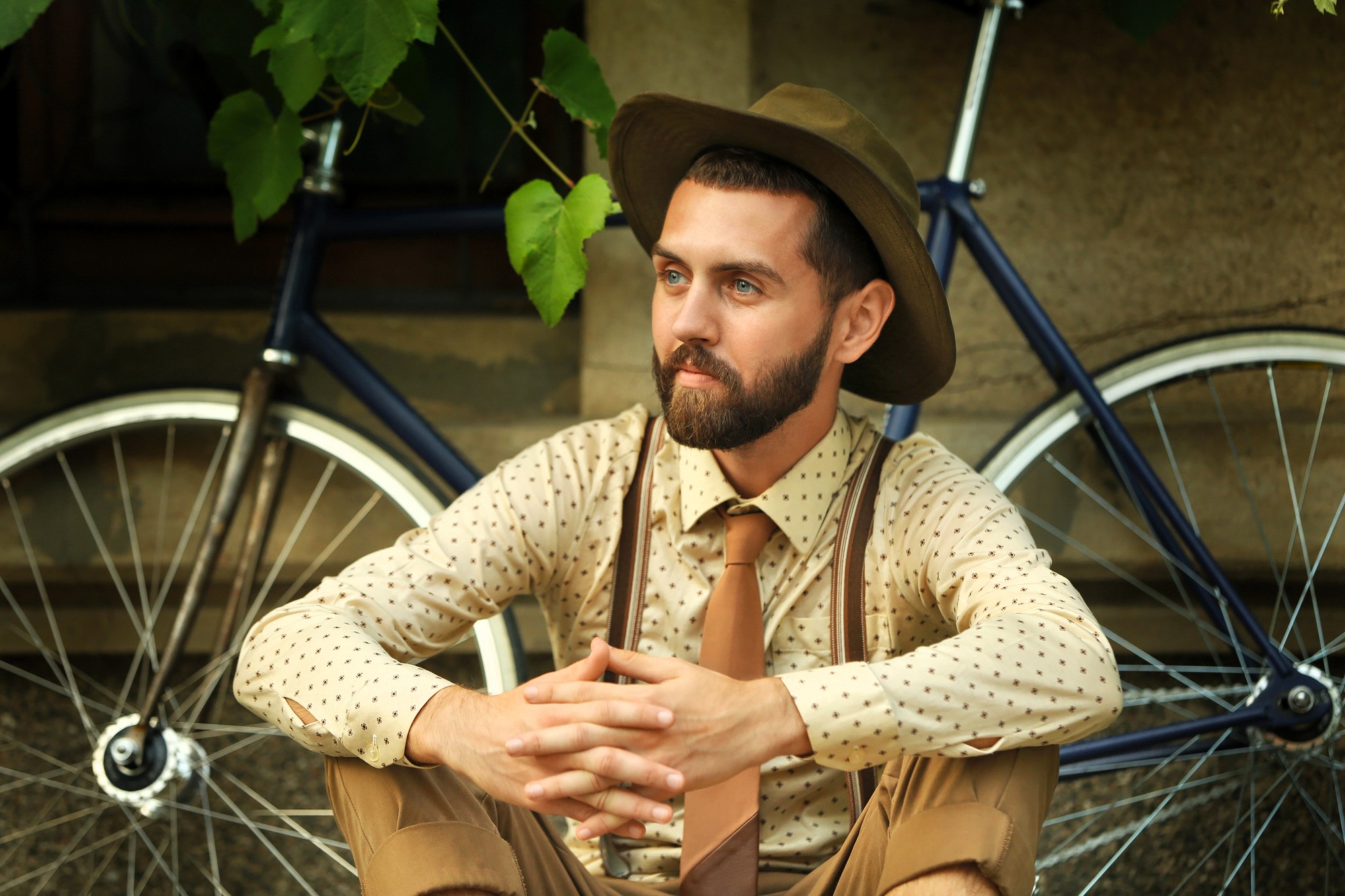 A bearded man in a polka-dot shirt, tie, suspenders, and wide-brimmed hat sits cross-legged outdoors, hands clasped, with a vintage bicycle and greenery in the background.