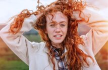 A young woman with long curly red hair holds her hands in her hair, smiling slightly. She wears a light jacket over a plaid shirt and stands outdoors with a natural, green landscape in the background.