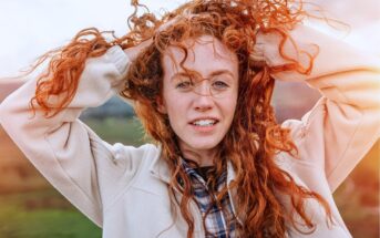 A young woman with long curly red hair holds her hands in her hair, smiling slightly. She wears a light jacket over a plaid shirt and stands outdoors with a natural, green landscape in the background.