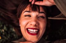 A person lying down, smiling widely with bright red lipstick and bold eyeliner, holding their bangs with one hand. The image captures a joyful, close-up expression.