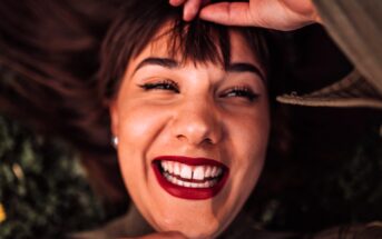 A person lying down, smiling widely with bright red lipstick and bold eyeliner, holding their bangs with one hand. The image captures a joyful, close-up expression.