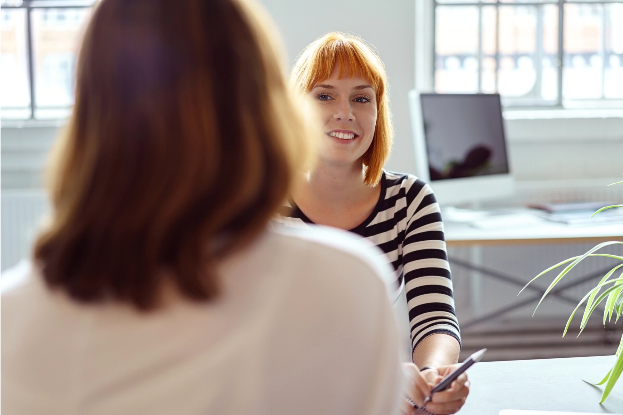 A woman with short red hair and a striped shirt smiles while holding a pen, sitting across a desk from another woman in an office with large windows and a computer in the background.