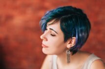 A person with short, wavy teal and blue hair and dangling earrings is shown in profile against a blurred brick wall, with eyes closed and a calm expression.