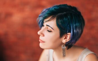 A person with short, wavy teal and blue hair and dangling earrings is shown in profile against a blurred brick wall, with eyes closed and a calm expression.