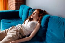 A young woman with long red hair lies on a bright blue sofa, eyes closed and face tense, appearing unwell or in pain. She wears a white top and beige pants, and sunlight streams in from the side.