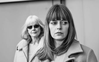 A younger woman with straight hair looks away pensively in the foreground, while an older woman wearing sunglasses stands behind her, both dressed in coats. The image is in black and white.
