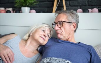 An older couple relaxes together on a bed, smiling and looking at each other affectionately. The woman rests her head on the man's shoulder. Cozy decor is visible in the background.