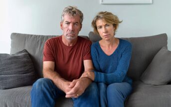 A middle-aged man and woman sit close together on a gray couch, looking seriously at the camera. The woman has her arm linked with the man's, and both appear concerned or worried. Neutral-colored pillows are in the background.