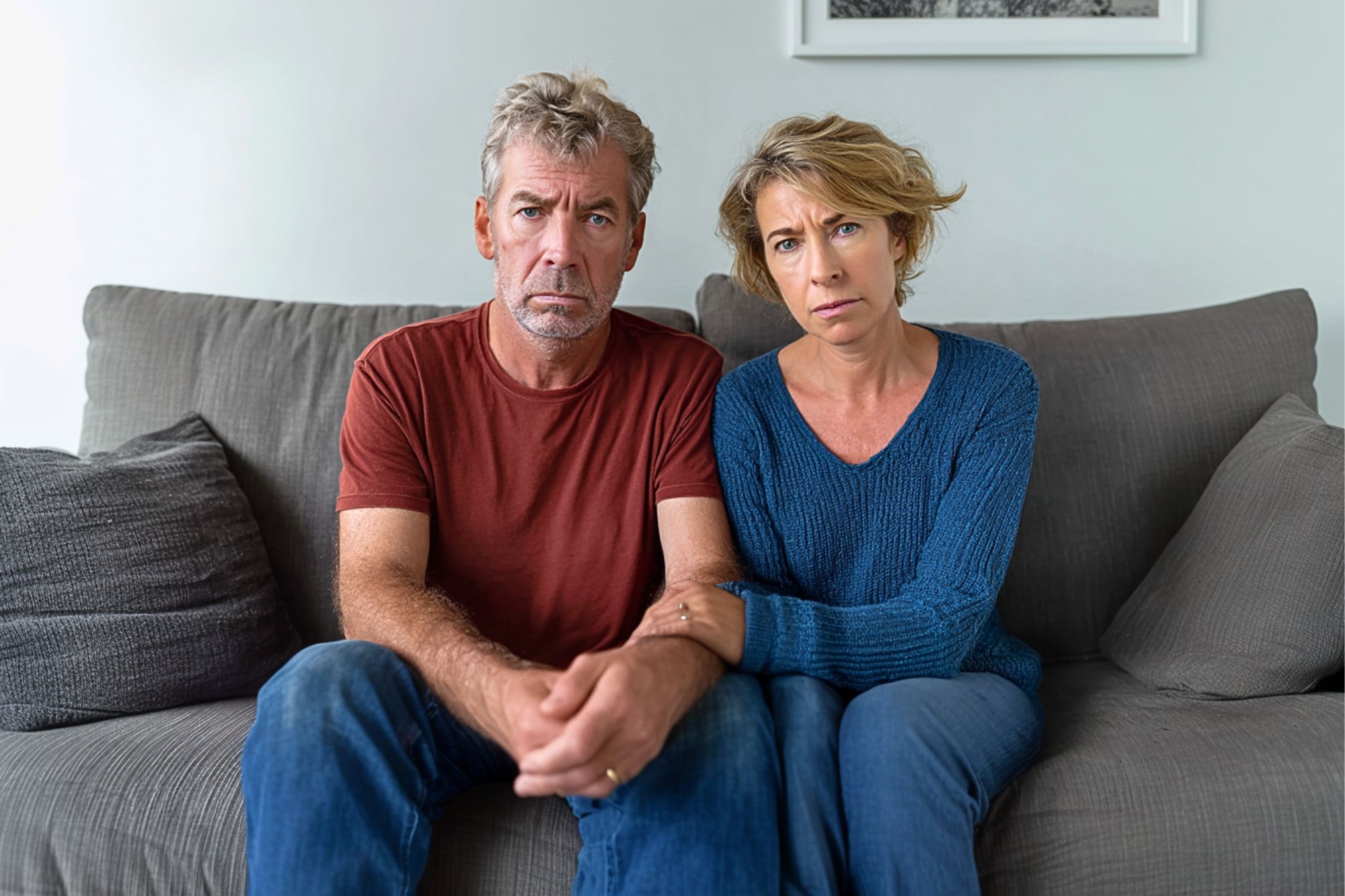 A middle-aged man and woman sit close together on a gray couch, looking seriously at the camera. The woman has her arm linked with the man's, and both appear concerned or worried. Neutral-colored pillows are in the background.