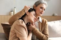 An older woman sits on a couch, gently embracing a younger woman who rests her head on the elder’s shoulder. Both appear thoughtful and comforted, sharing an intimate, supportive moment in a cozy, softly lit room.