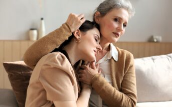 An older woman sits on a couch, gently embracing a younger woman who rests her head on the elder’s shoulder. Both appear thoughtful and comforted, sharing an intimate, supportive moment in a cozy, softly lit room.