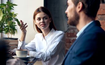 A woman in a white shirt sits at a café table with a coffee cup, gesturing and speaking to a man in a blue jacket. They appear to be having a serious conversation near a window with natural light.