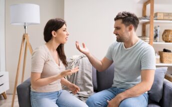 A woman and man sit on a couch in a living room, facing each other and appearing to have an intense discussion or argument, with expressive hand gestures and concerned facial expressions.