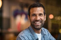 A man with short dark hair and a beard smiles warmly at the camera, wearing a blue patterned shirt. The background is softly blurred, suggesting an indoor setting with ambient lighting.