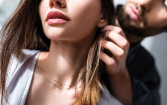 A close-up of a woman with loose hair and red lipstick as a man gently touches her hair, creating an intimate and romantic atmosphere. Both faces are partially visible, and the lighting is soft.