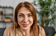 A middle-aged woman with long reddish-brown hair and blue eyes smiles warmly at the camera. She is indoors, with a blurred green plant and shelves visible in the background.