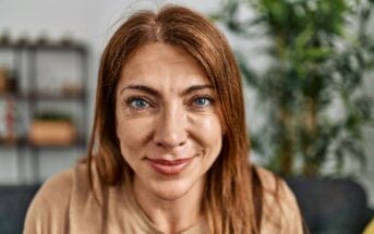 A middle-aged woman with long reddish-brown hair and blue eyes smiles warmly at the camera. She is indoors, with a blurred green plant and shelves visible in the background.