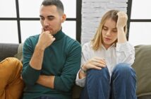 A man and a woman sit on a couch looking upset and distant. The man rests his chin on his hand, while the woman holds her forehead and looks down. Both appear to be deep in thought or troubled.