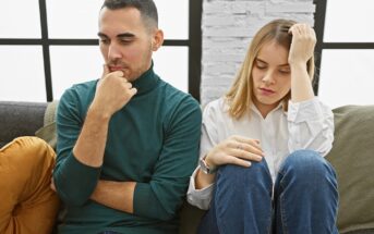 A man and a woman sit on a couch looking upset and distant. The man rests his chin on his hand, while the woman holds her forehead and looks down. Both appear to be deep in thought or troubled.