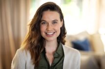 A woman with long brown hair and a light complexion smiles at the camera. She is wearing a green shirt and a light-colored cardigan, with a softly blurred indoor background behind her.