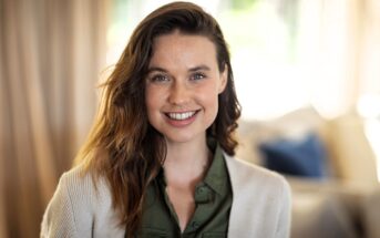 A woman with long brown hair and a light complexion smiles at the camera. She is wearing a green shirt and a light-colored cardigan, with a softly blurred indoor background behind her.