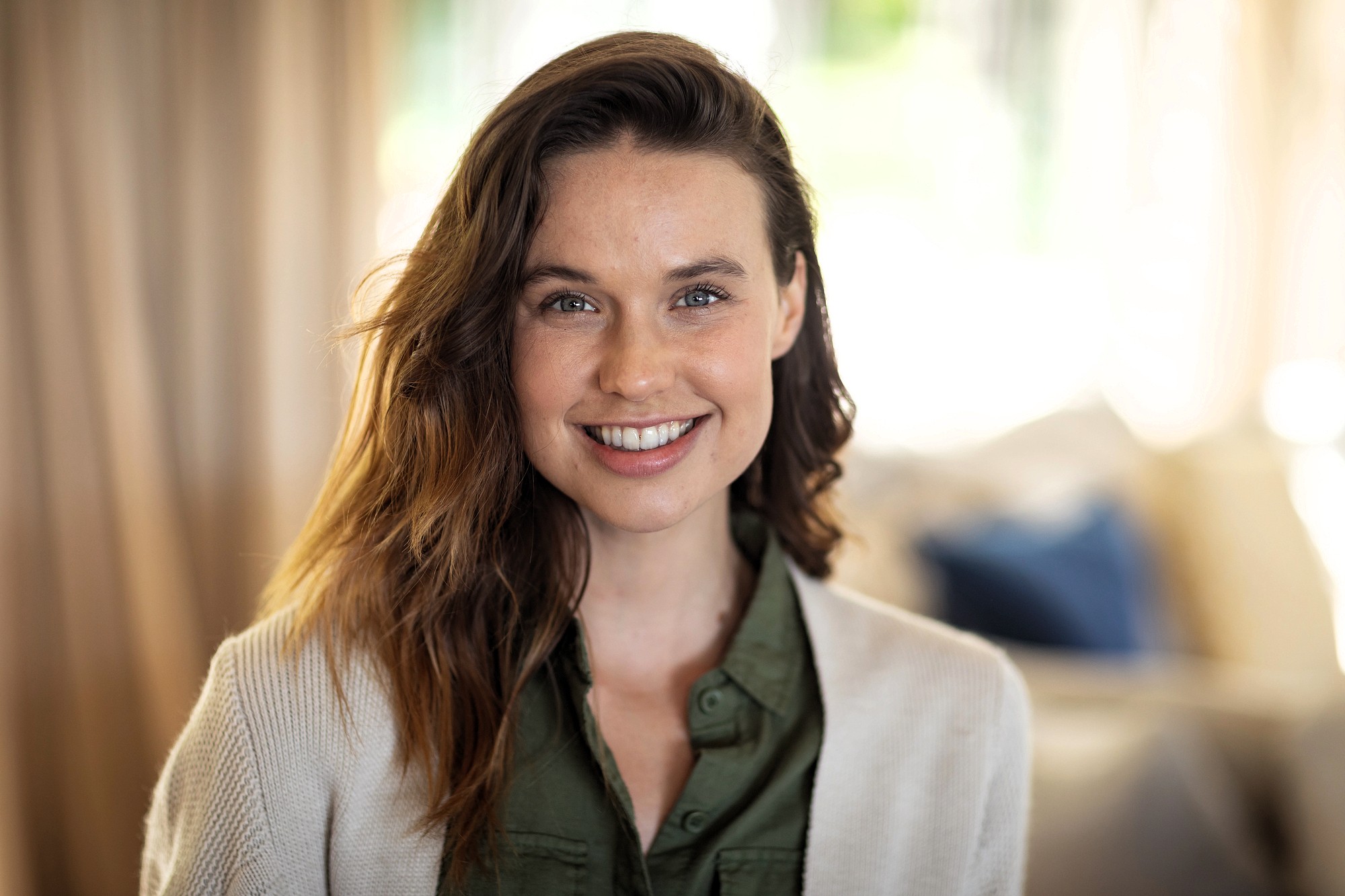 A woman with long brown hair and a light complexion smiles at the camera. She is wearing a green shirt and a light-colored cardigan, with a softly blurred indoor background behind her.