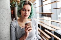 Young woman with short, wavy turquoise hair and a gray shirt holds a takeaway coffee cup while leaning against a concrete wall in a modern, sunlit cafe.