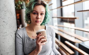 Young woman with short, wavy turquoise hair and a gray shirt holds a takeaway coffee cup while leaning against a concrete wall in a modern, sunlit cafe.
