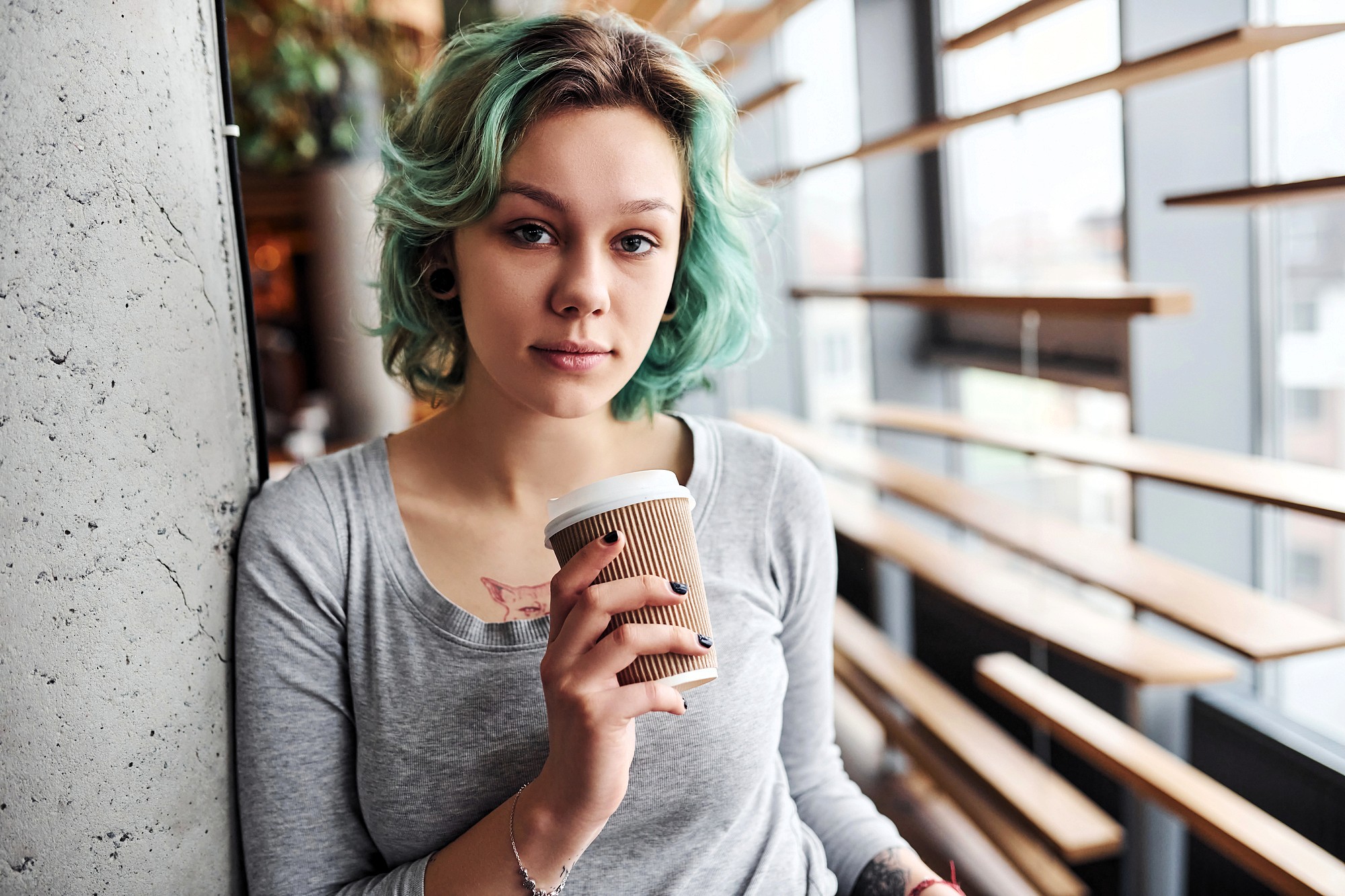 Young woman with short, wavy turquoise hair and a gray shirt holds a takeaway coffee cup while leaning against a concrete wall in a modern, sunlit cafe.