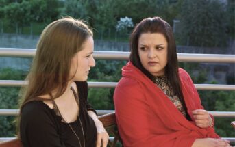 Two women sit outdoors on a bench, facing each other. One woman wears black and looks calm, while the other, draped in a red shawl, looks at her with a serious, concerned expression. Green trees are visible in the background.