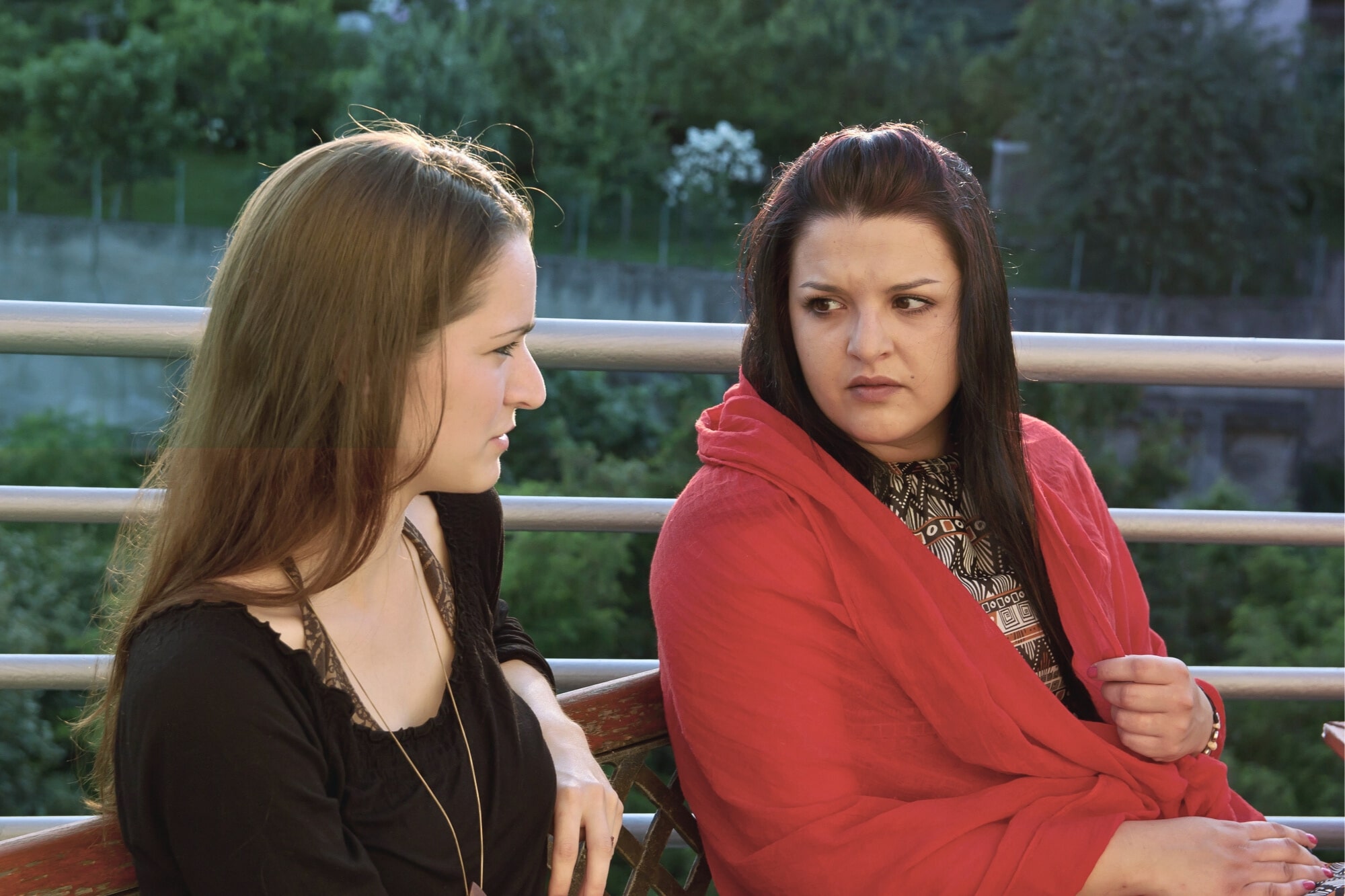 Two women sit outdoors on a bench, facing each other. One woman wears black and looks calm, while the other, draped in a red shawl, looks at her with a serious, concerned expression. Green trees are visible in the background.