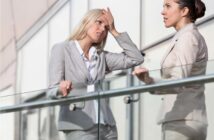 Two women in business attire standing outside by a glass railing, having a conversation; one woman looks stressed with her hand on her forehead, while the other listens attentively.