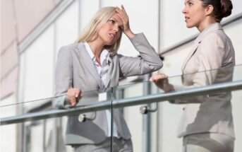 Two women in business attire standing outside by a glass railing, having a conversation; one woman looks stressed with her hand on her forehead, while the other listens attentively.