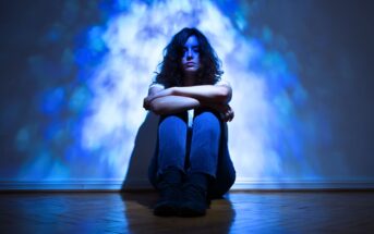A person with wavy hair sits on the floor against a wall, arms wrapped around their knees, with blue and white light patterns projected behind them, creating a dramatic and moody atmosphere.