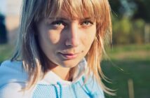 A young person with blonde hair and green eyes looks at the camera, standing outdoors in sunlight, wearing a light blue hoodie. The background is blurred greenery.