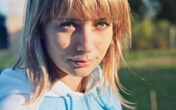 A young person with blonde hair and green eyes looks at the camera, standing outdoors in sunlight, wearing a light blue hoodie. The background is blurred greenery.