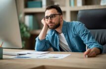 A man with glasses and a beard sits at a desk, resting his chin on his hand and looking thoughtfully at a computer screen. Papers, a pen, and office supplies are spread out in front of him.