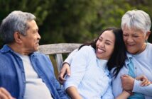 Three people sit close together on a wooden bench outdoors, smiling and laughing. An older man and woman embrace a young woman between them, all appearing happy and relaxed. Greenery is visible in the background.