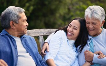 Three people sit close together on a wooden bench outdoors, smiling and laughing. An older man and woman embrace a young woman between them, all appearing happy and relaxed. Greenery is visible in the background.
