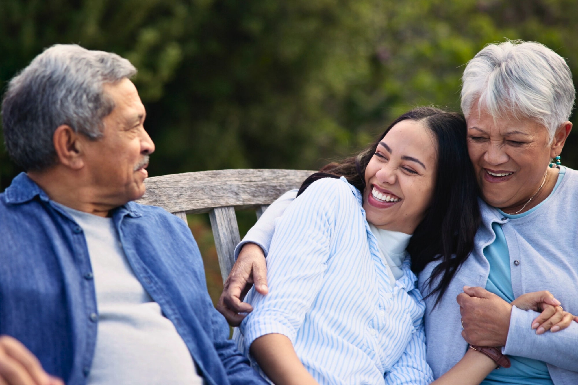 Three people sit close together on a wooden bench outdoors, smiling and laughing. An older man and woman embrace a young woman between them, all appearing happy and relaxed. Greenery is visible in the background.