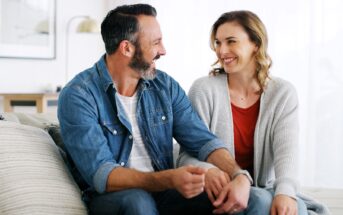 A smiling man and woman sit closely together on a couch, holding hands and looking at each other warmly in a cozy, well-lit living room.